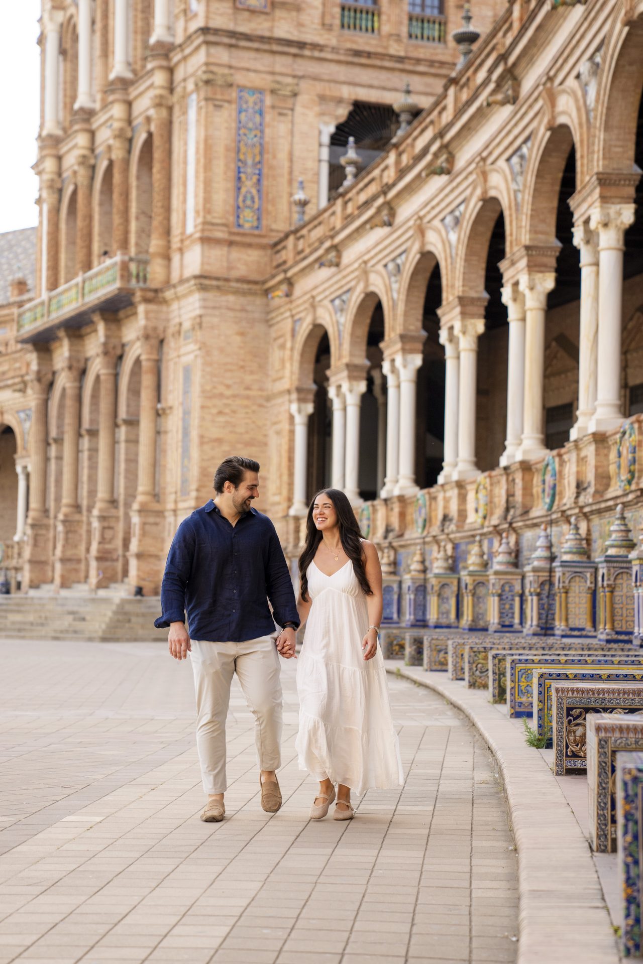 Couple walking hand in hand past the tiled alcoves of Plaza de España, Seville